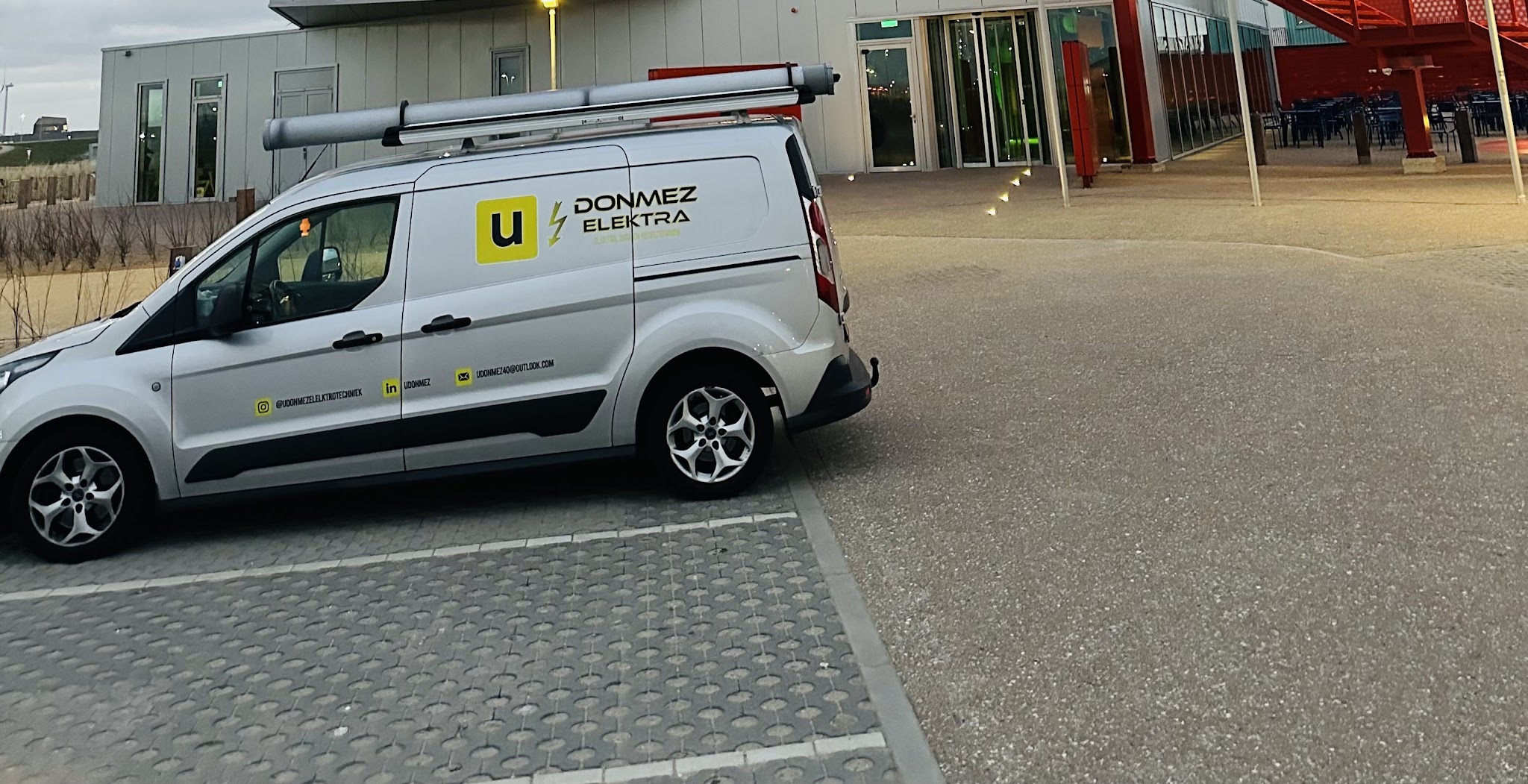 De zilvergrijze bedrijfsbus van Donmez Elektra met het gele u-logo, geparkeerd voor een gebouw
